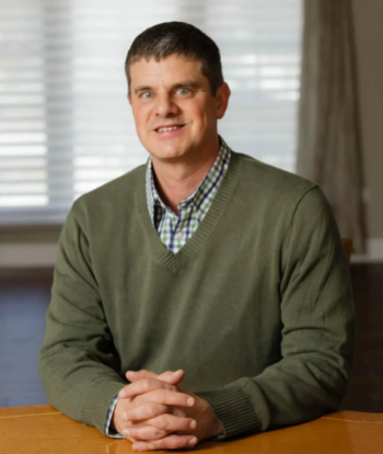 Mike Wasilewski in a dark green sweater and check shirt, sitting with clasped hands